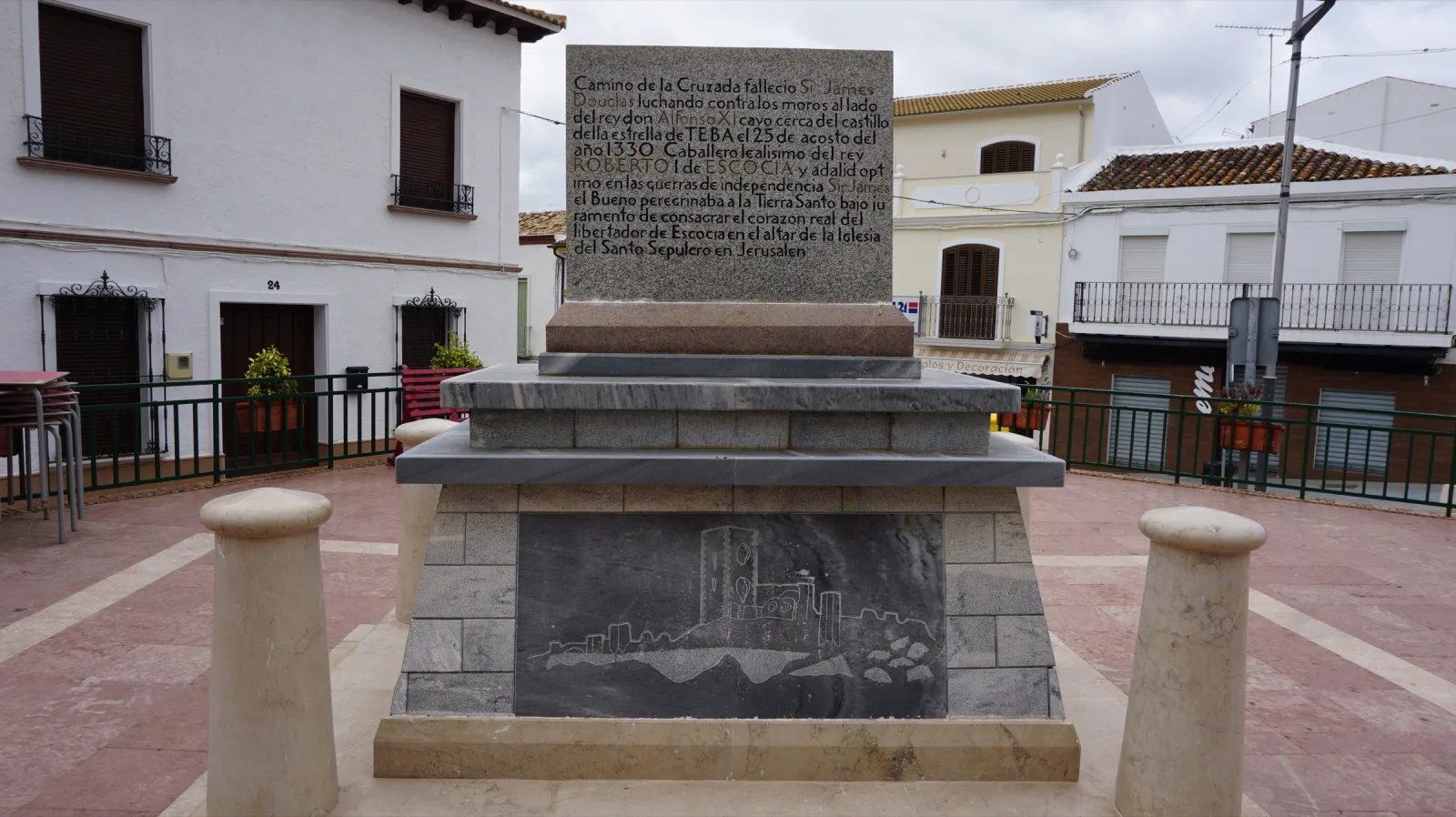 Monumento de piedra en una plaza de Teba: una losa vertical con una inscripción sobre un pedestal escalonado con un relieve de un castillo, rodeado de pequeños bolardos, con casas blancas al fondo.