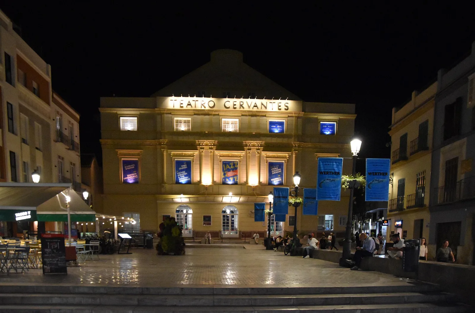 Fachada iluminada del Teatro Cervantes de Málaga, de noche, con banderolas azules en la plaza.