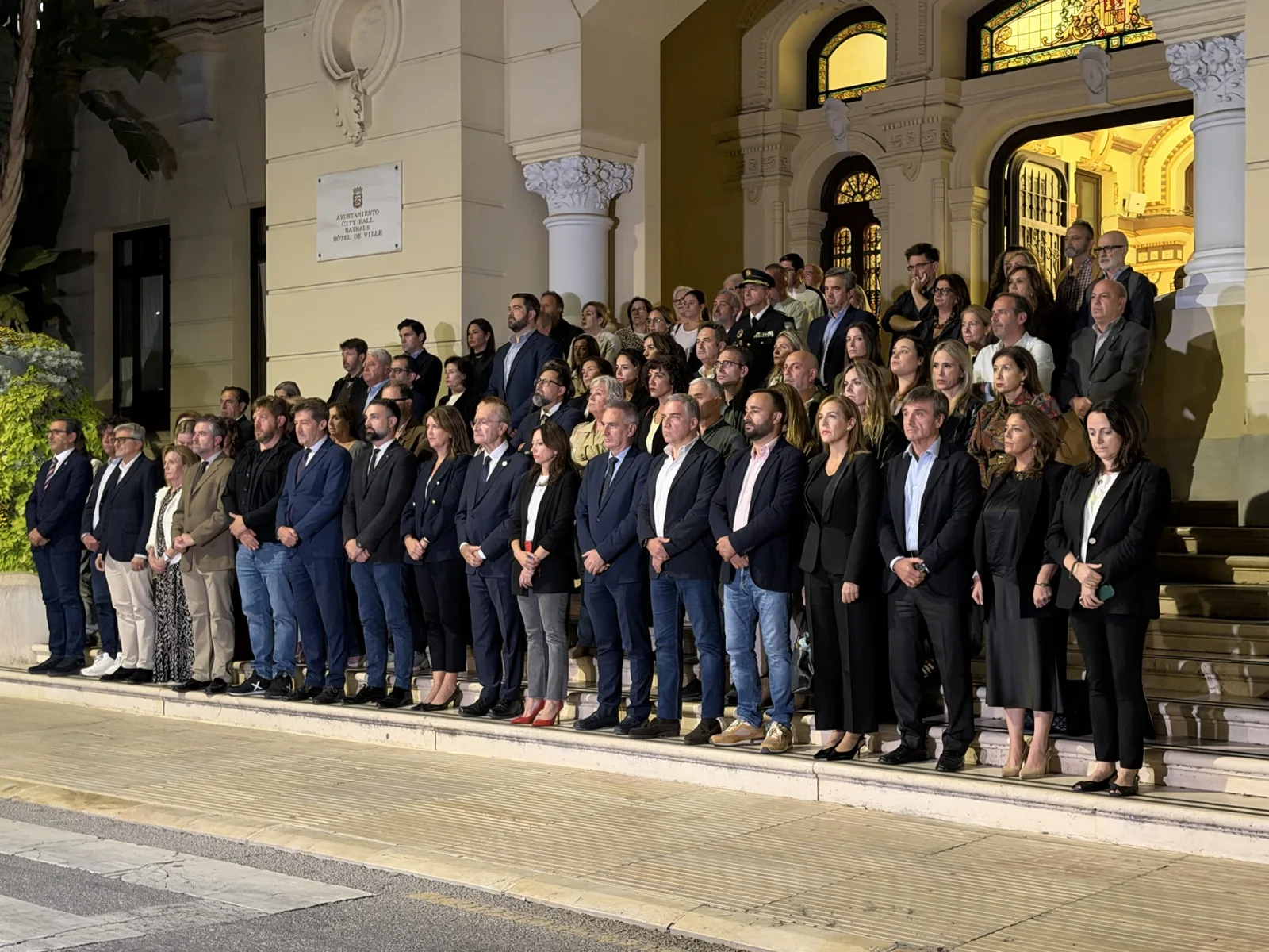 Decenas de personas guardan un minuto de silencio de pie en la escalinata del Ayuntamiento de Málaga, de noche.