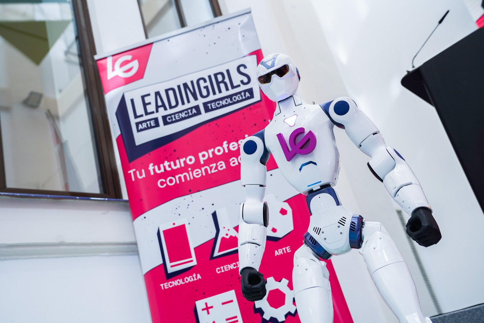 Un robot humanoide blanco y negro de pie junto a un cartel rosa y blanco con el texto Leadingirls y las palabras arte, ciencia y tecnología.