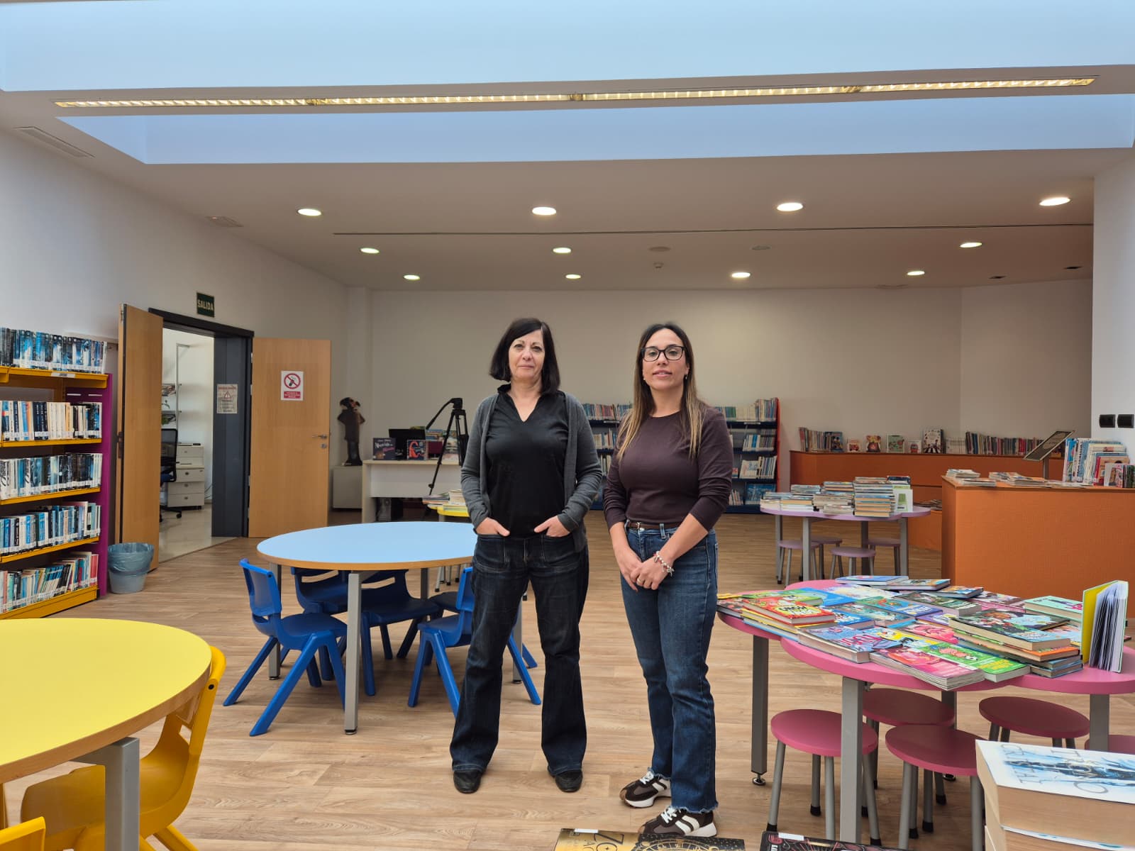 Dos mujeres de pie en una sala amplia con mesas redondas, sillas de colores y estanterías con libros.