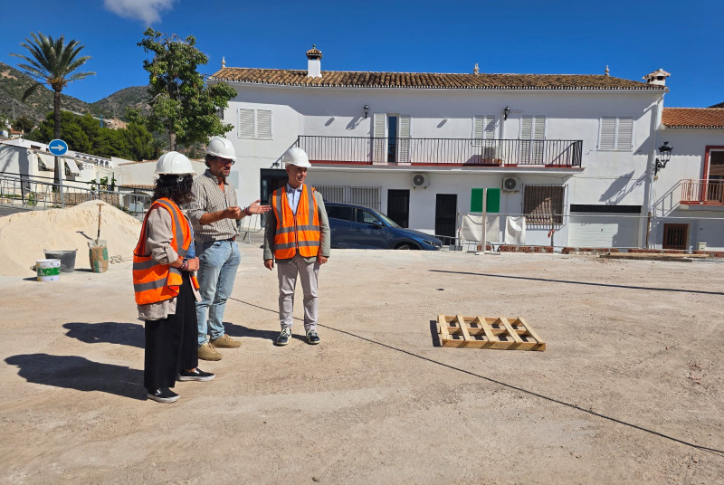 Tres personas con casco y chaleco reflectante de obra charlan en una explanada de tierra frente a un edificio blanco con balcón.