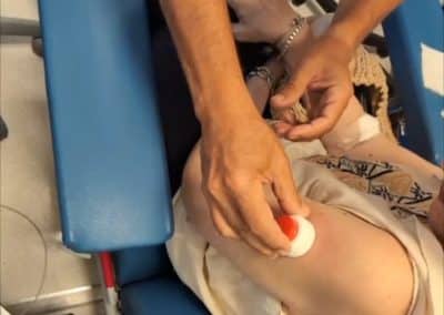 Unas manos colocan un algodón con líquido rojo en el brazo de una persona sentada en una silla azul.