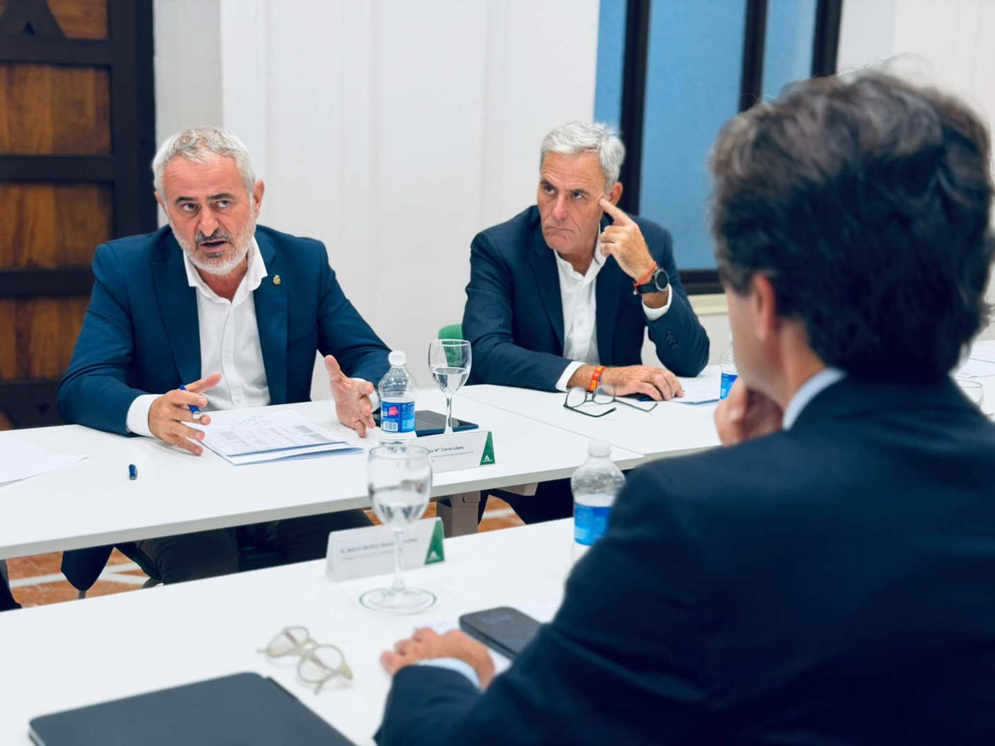 Tres hombres trajeados conversan sentados alrededor de una mesa blanca con documentos, botellas de agua y vasos.