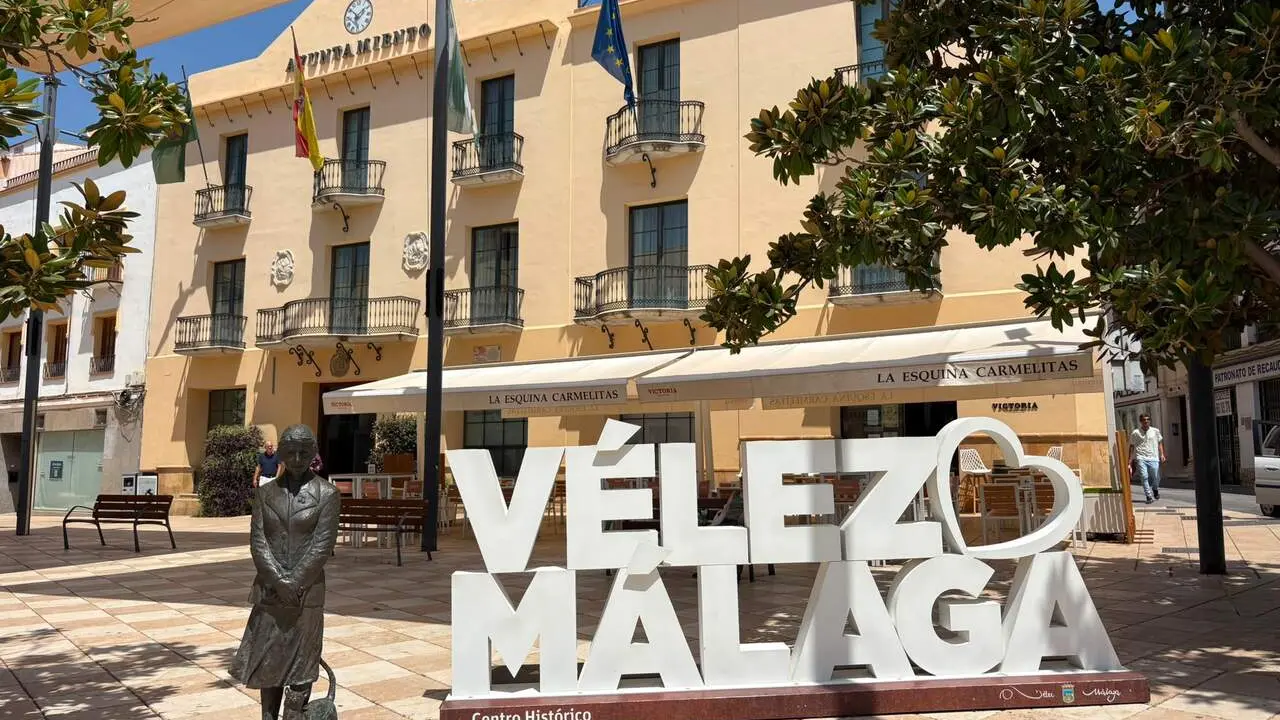 Letras gigantes blancas con VÉLEZ MÁLAGA en una plaza, ante un edificio amarillo con banderas y una estatua.