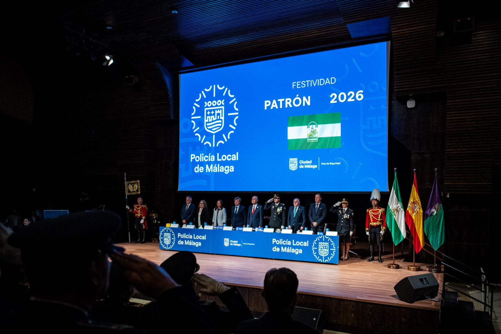 Una fila de personas de pie en un escenario ante una gran pantalla azul con el texto Policía Local de Málaga y FESTIVIDAD PATRÓN 2026, junto a banderas.