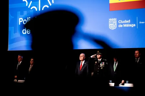 Varias personas trajeadas de pie ante una pantalla azul con el escudo de Málaga, una de ellas uniformada y saludando.