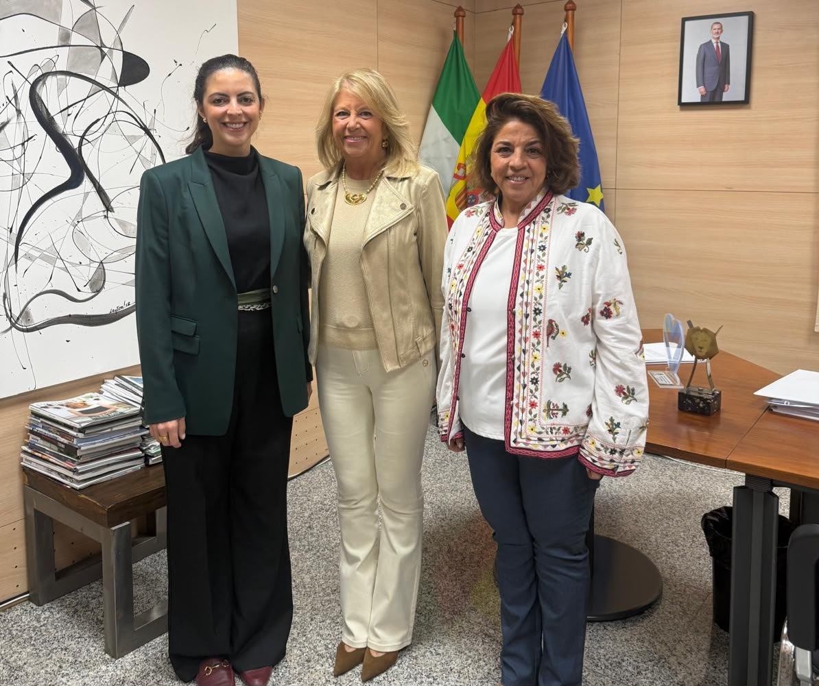 Tres mujeres de pie sonriendo en una sala con banderas, un cuadro abstracto y un retrato enmarcado en la pared.