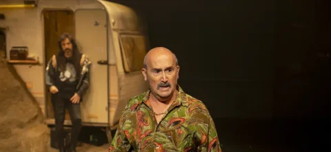 Un hombre calvo con bigote y camisa estampada habla en primer plano ante una caravana, con otra persona al fondo.