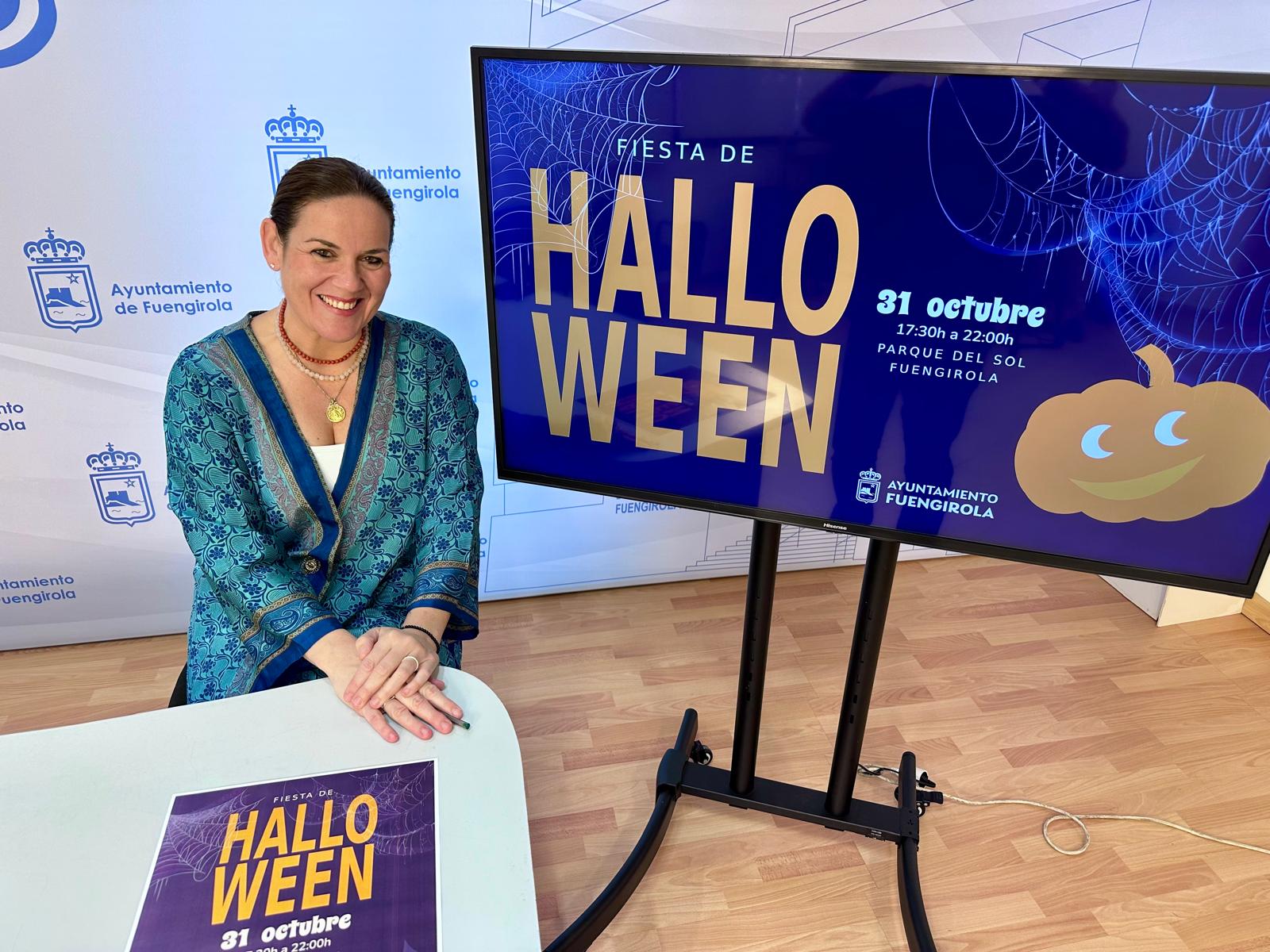 Una mujer sonriente de chaqueta azul posa junto a una pantalla que anuncia una fiesta de Halloween el 31 de octubre en el Parque del Sol.