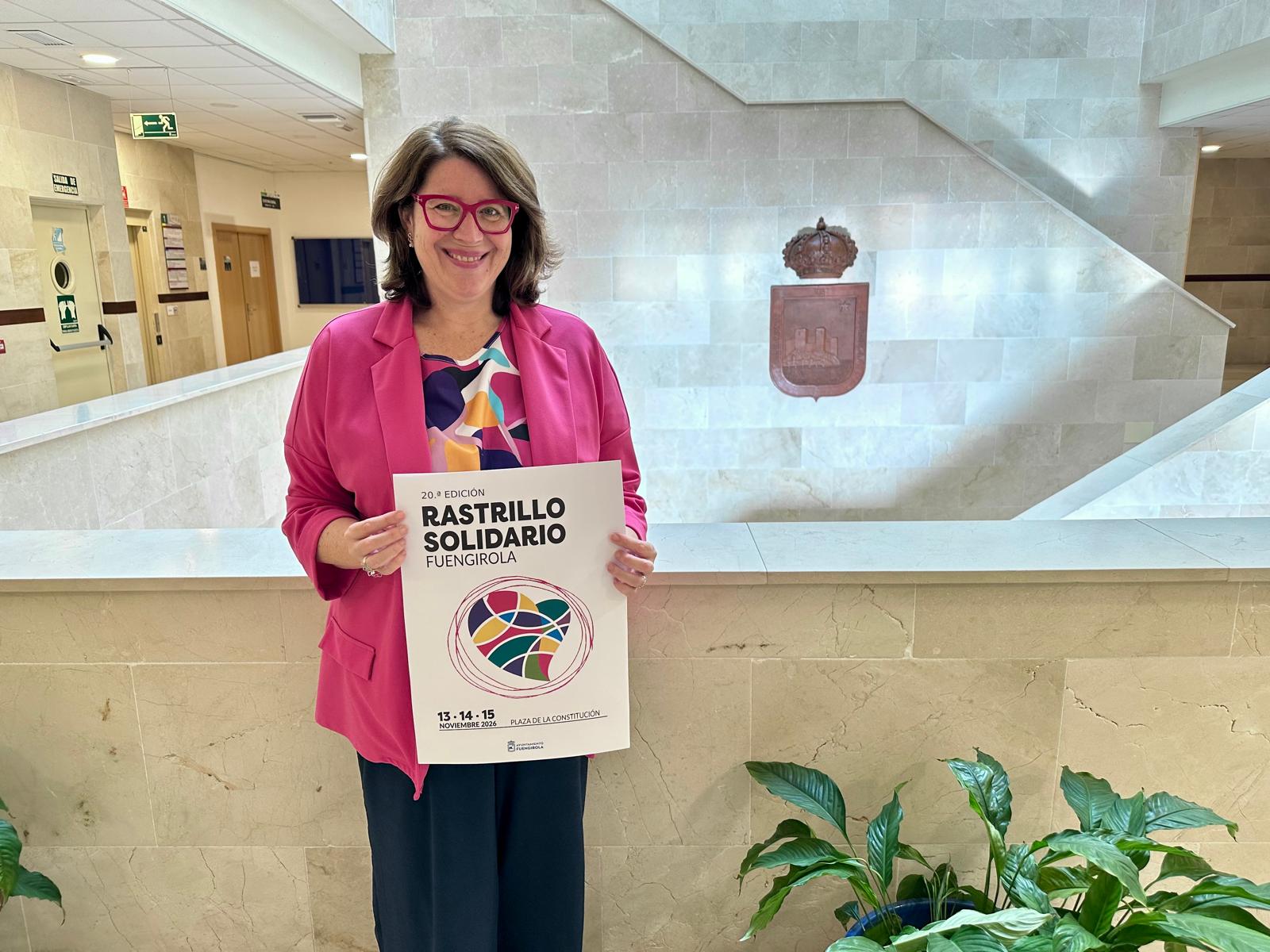 Una mujer con gafas y chaqueta rosa sostiene un cartel del XX Rastrillo Solidario en un interior con escaleras y un escudo en la pared.