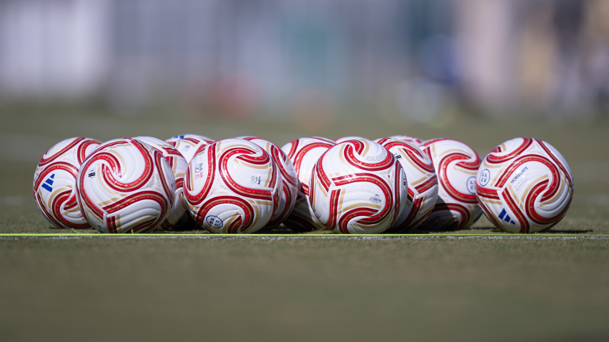Varios balones de fútbol blancos con dibujos rojos y dorados alineados sobre el césped de un campo.
