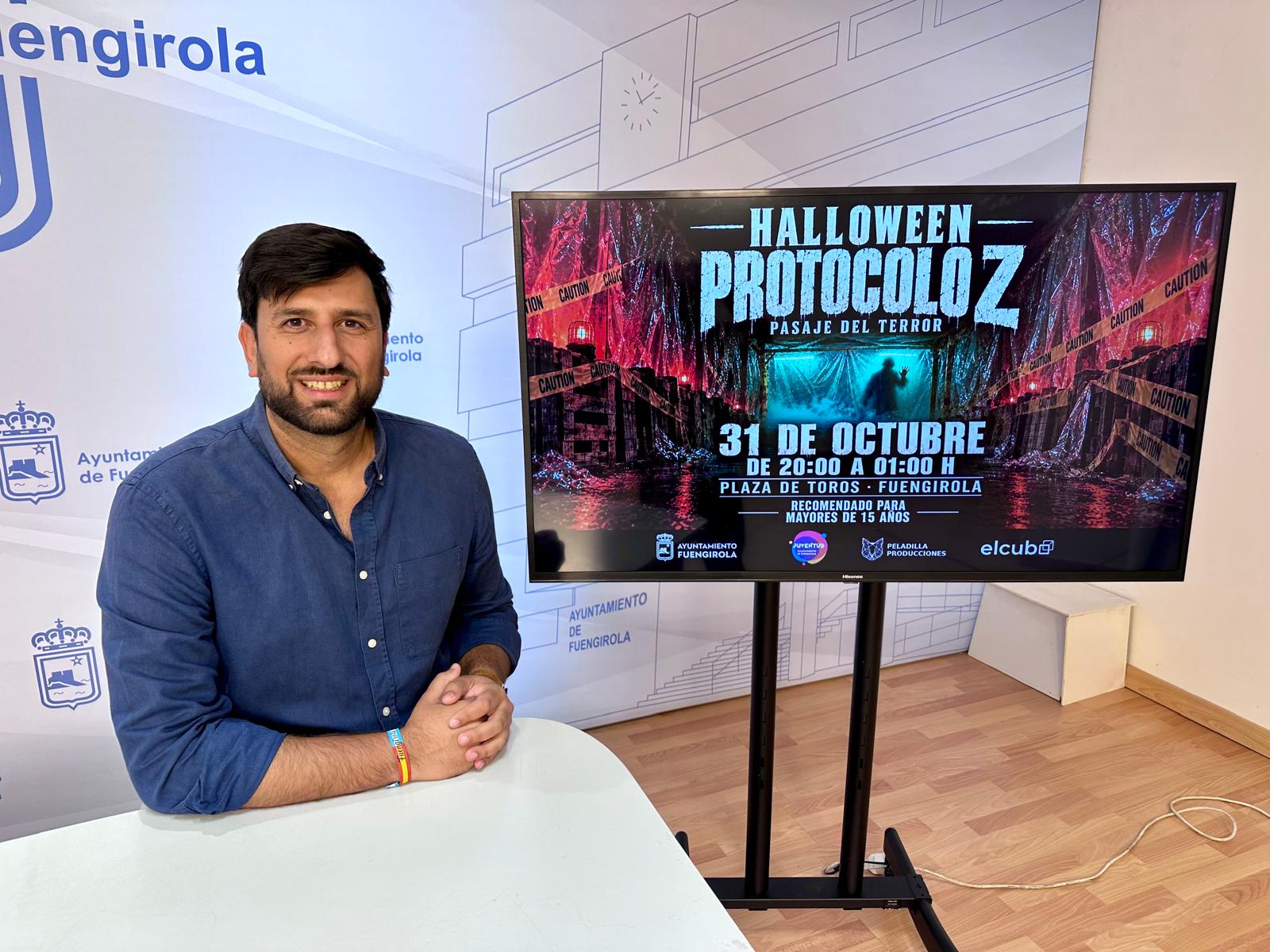 Un hombre sonriente con camisa azul posa junto a una pantalla que anuncia un pasaje del terror de Halloween.