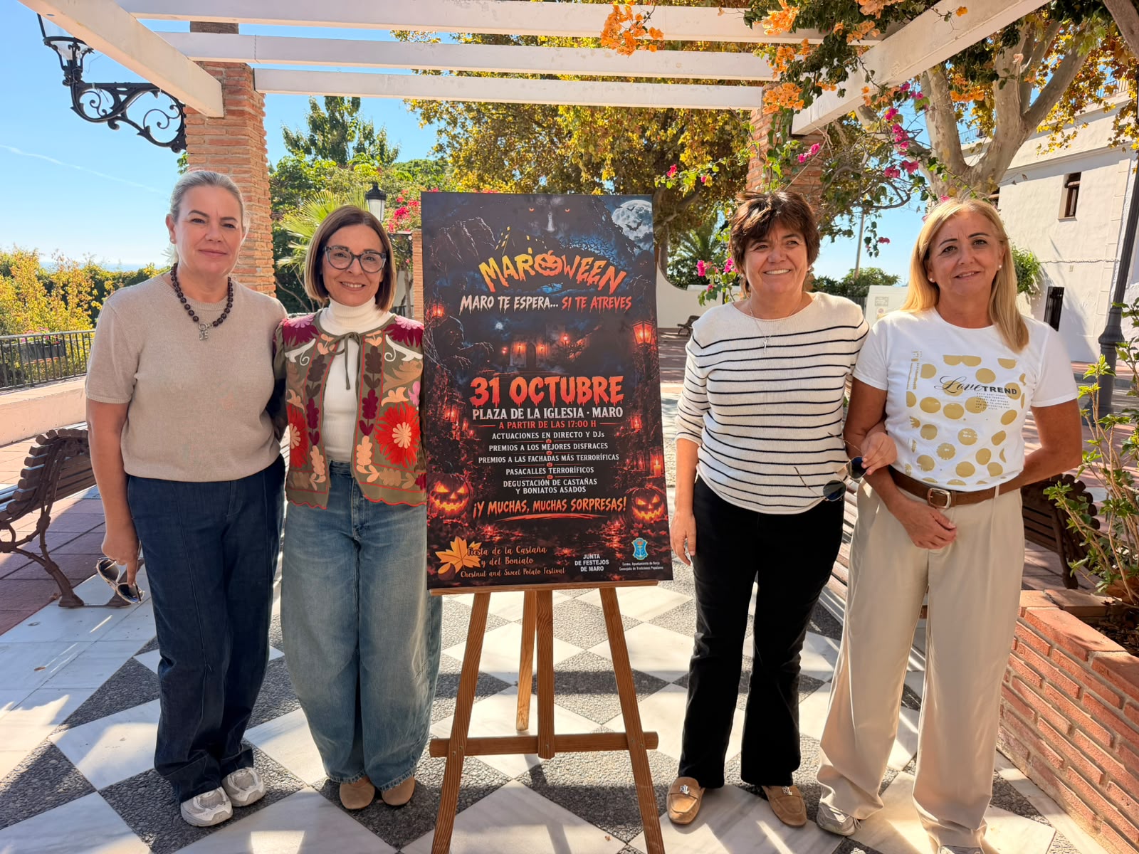 Cuatro mujeres de pie junto a un cartel de Maroween sobre un caballete en una plaza exterior.