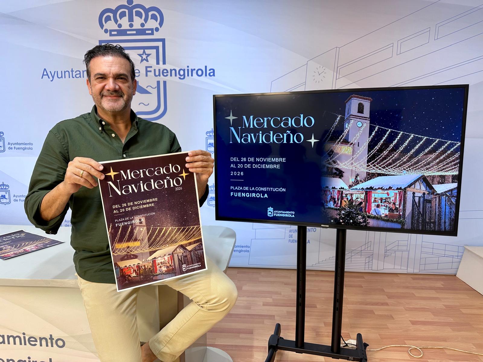 Un hombre de pie sostiene un cartel del Mercado Navideño junto a una pantalla con el mismo cartel y un logotipo municipal de fondo.