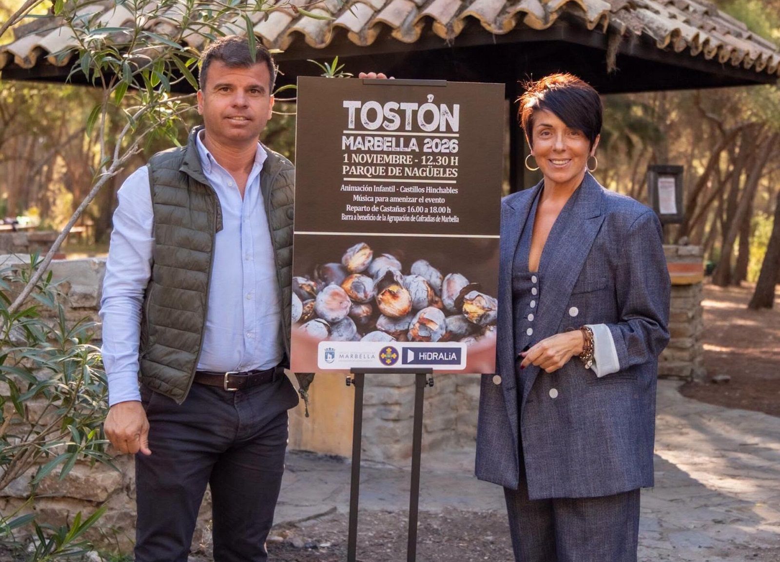 Un hombre y una mujer posan junto a un cartel sobre el Tostón de Marbella con castañas, bajo una estructura de techo.