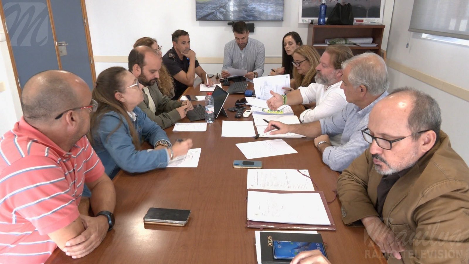 Unas diez personas sentadas alrededor de una mesa de reuniones con documentos, portátiles y móviles.