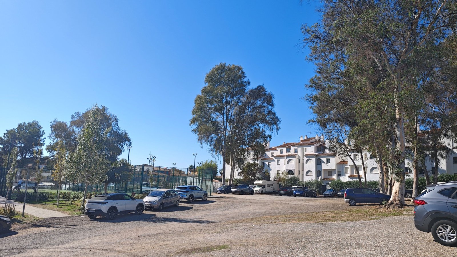 Una explanada de tierra con coches aparcados junto a árboles altos, una pista deportiva vallada y edificios blancos al fondo bajo un cielo despejado.