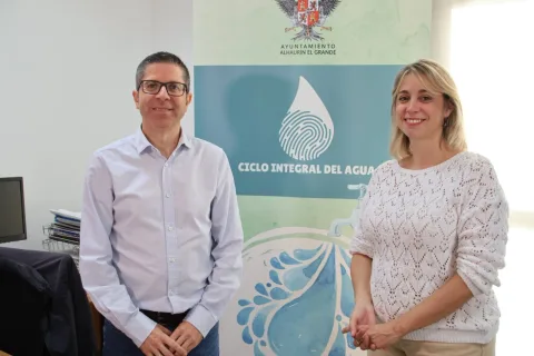 Un hombre con gafas y camisa azul y una mujer con jersey blanco de calado posan sonrientes delante de un cartel sobre el ciclo integral del agua.