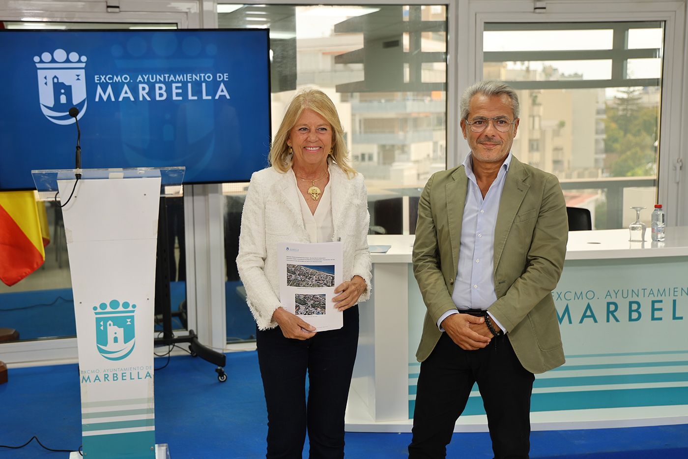Un hombre y una mujer de pie sostienen un documento con dos fotos aéreas ante un atril y una pantalla con el escudo del Ayuntamiento de Marbella.