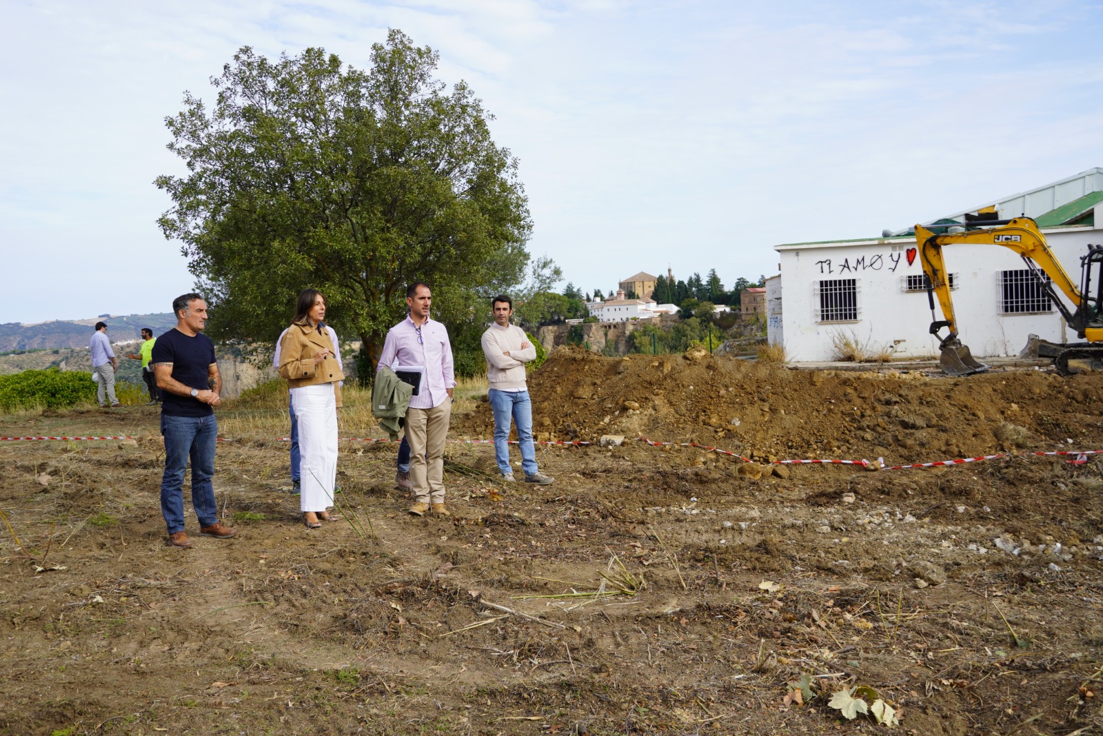 Cuatro personas de pie en un terreno de tierra removida junto a una excavadora amarilla y un edificio blanco, con un pueblo al fondo.
