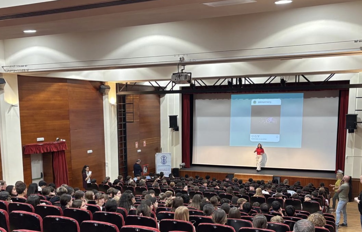 Una mujer habla en el escenario de un salón de actos ante un público joven sentado en butacas, con una pantalla de proyección detrás.