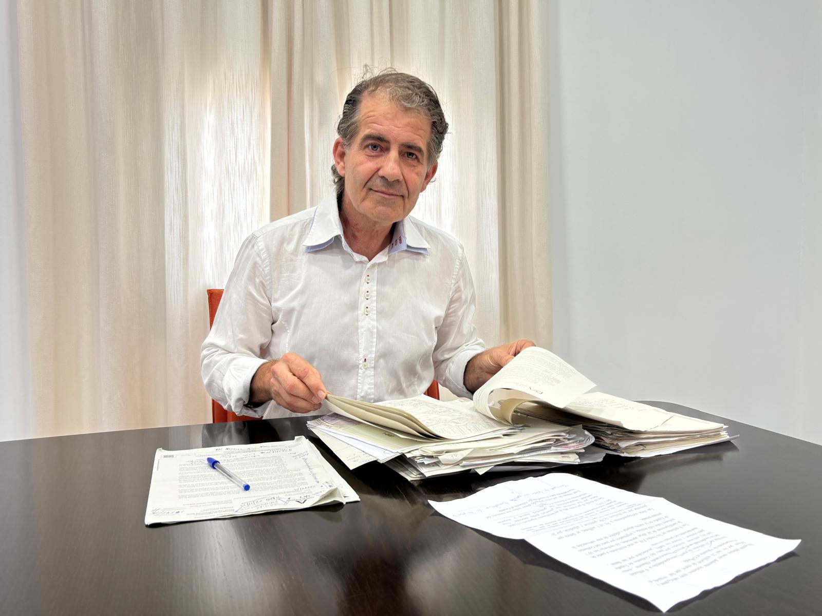 Un hombre de pelo canoso, con camisa blanca, sentado ante una mesa con montones de documentos y un bolígrafo.
