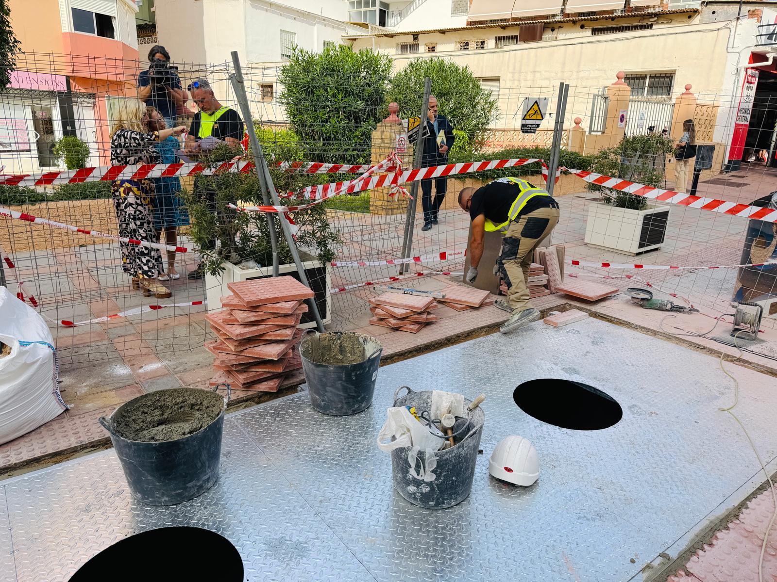 Un operario agachado coloca baldosas junto a pilas de losas, cubos de cemento y una valla metálica en una obra urbana.