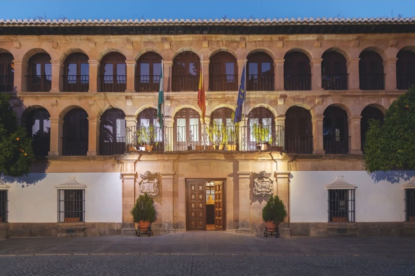 Fachada de piedra con dos plantas de arcos y balcones, puerta central de madera, escudos y tres banderas, al anochecer.