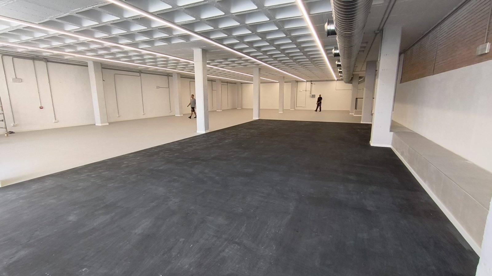 Nave diáfana vacía con suelo oscuro, paredes y pilares blancos, techo reticular con luces lineales y dos personas al fondo.
