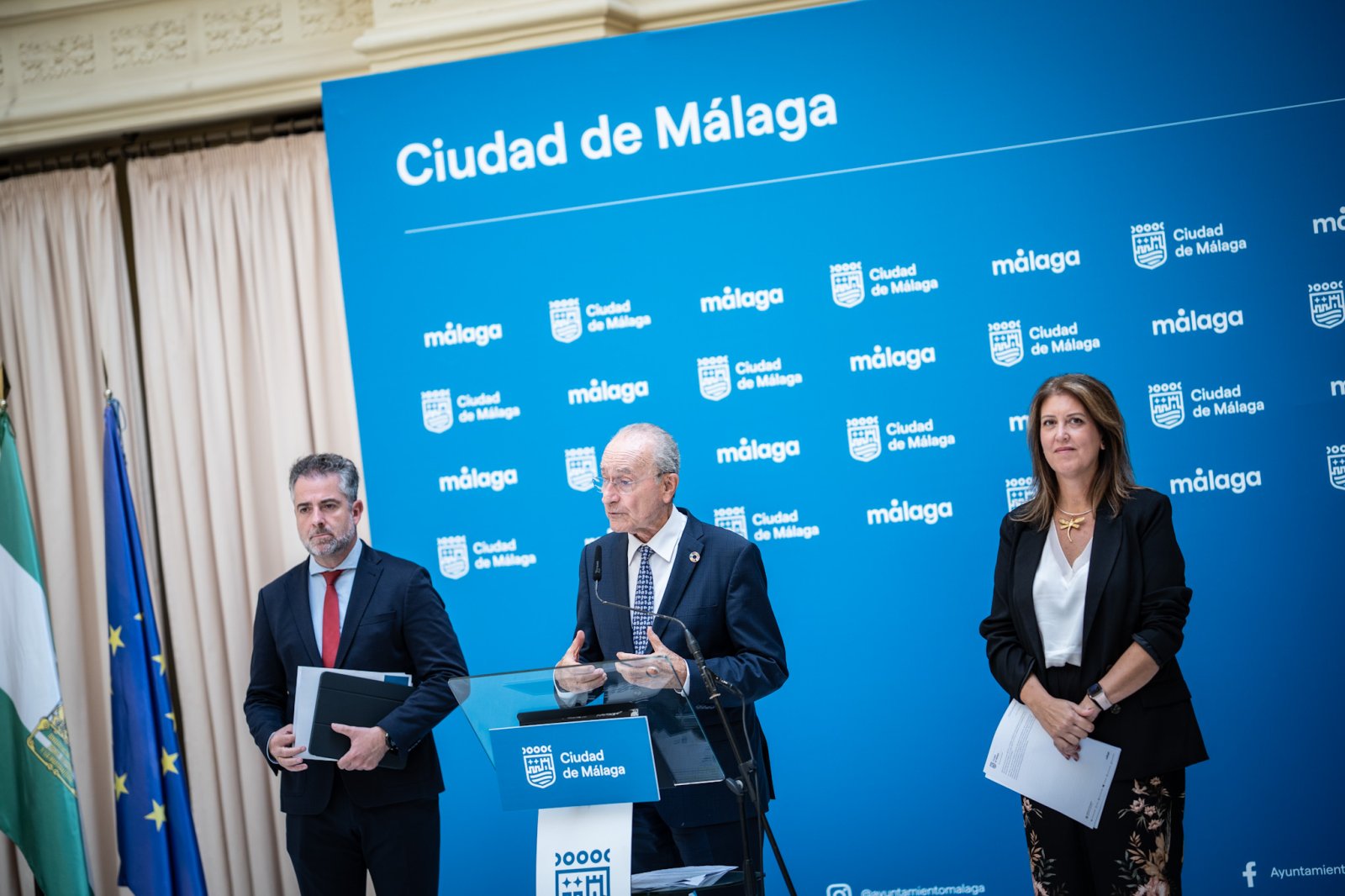 Tres personas trajeadas en un atril ante un panel azul con el texto Ciudad de Málaga y banderas.