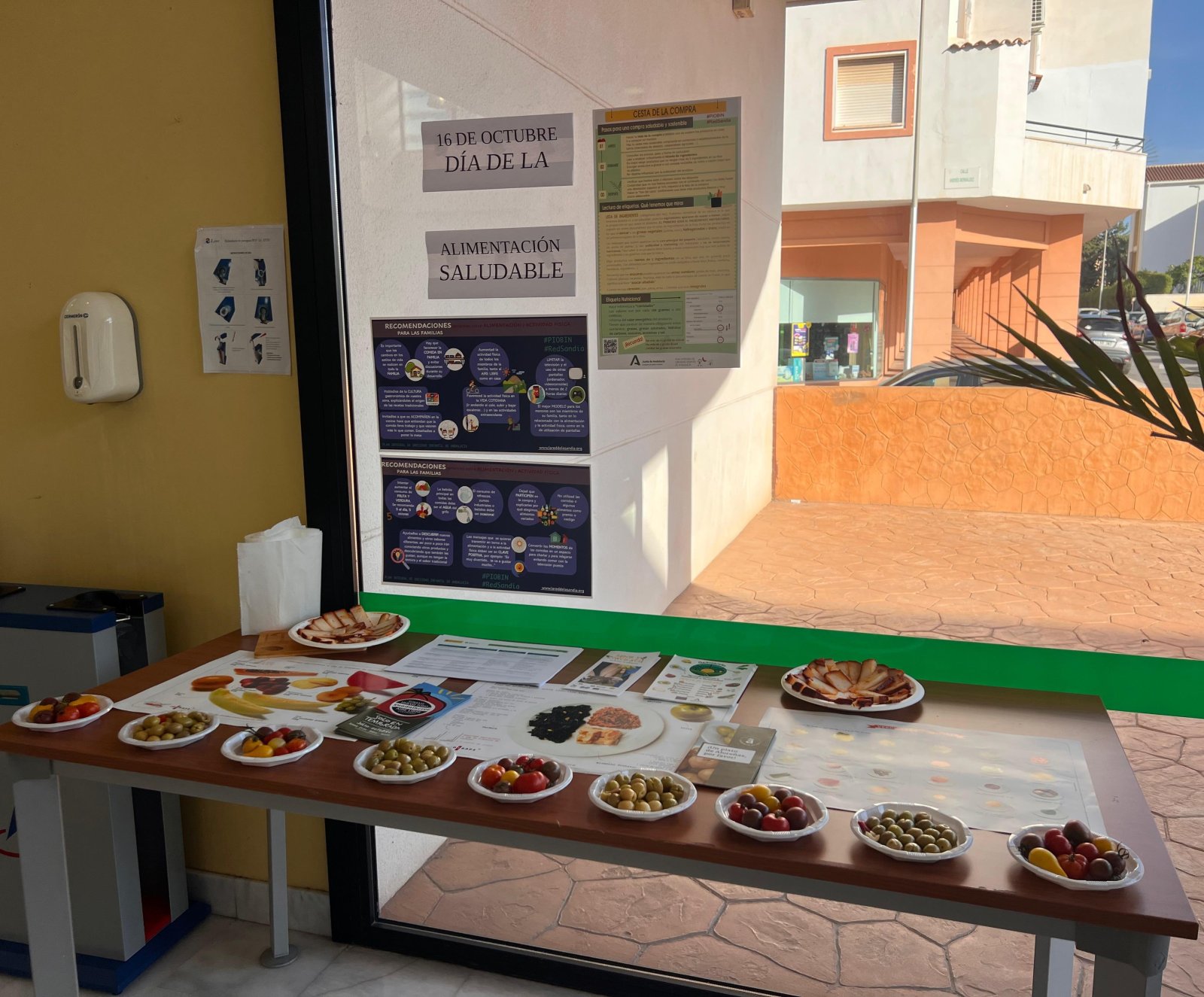 Mesa con platos de aceitunas, fruta y embutido junto a carteles sobre alimentación saludable en un centro de salud.