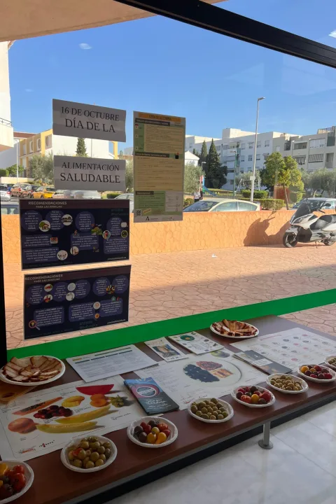Carteles sobre alimentación saludable y platos con aceitunas, tomates y fruta en una mesa junto a una ventana.
