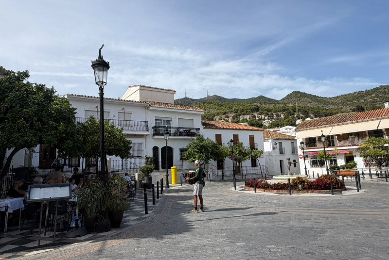Plaza empedrada con farolas, terrazas de bar, un hombre paseando y casas blancas con montañas al fondo.