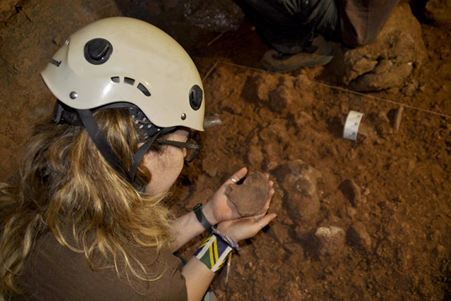 Una persona con casco blanco y gafas sostiene un fragmento de tierra en una excavación, junto a restos óseos y una etiqueta.