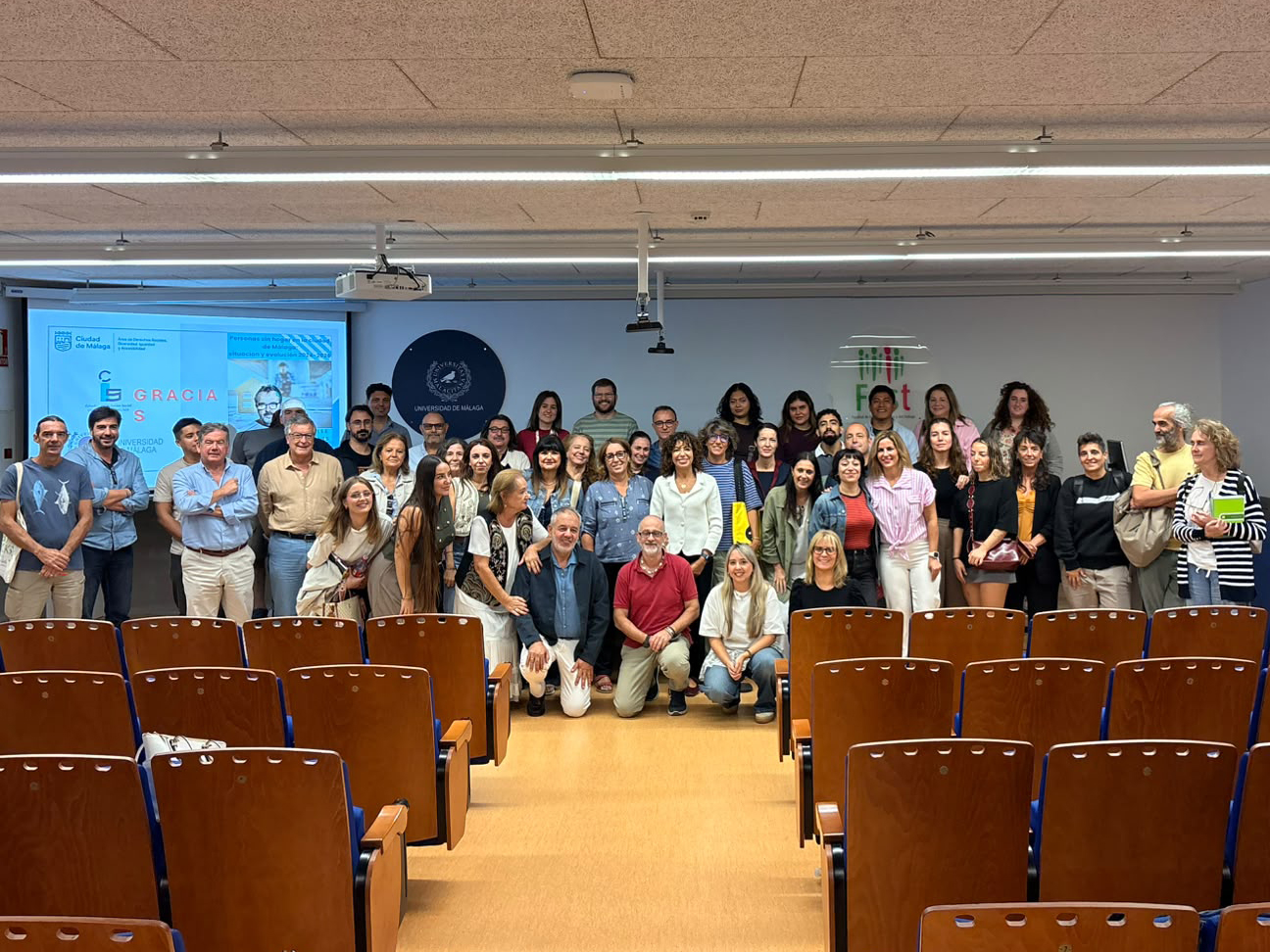 Un grupo de unas cuarenta personas posa de pie y agachadas en una sala con butacas de madera, pantalla y logotipos de la Universidad de Málaga.