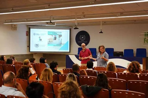 Dos personas de pie ante una pantalla de proyección en un aula con público sentado en butacas naranjas.