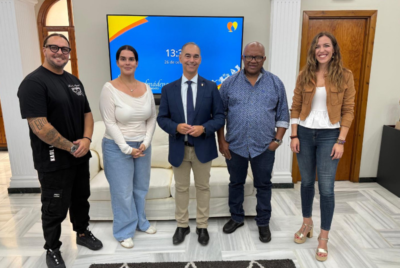 Cinco personas de pie posan sonrientes en una sala con pantalla azul al fondo, sofá blanco y suelo de mármol.