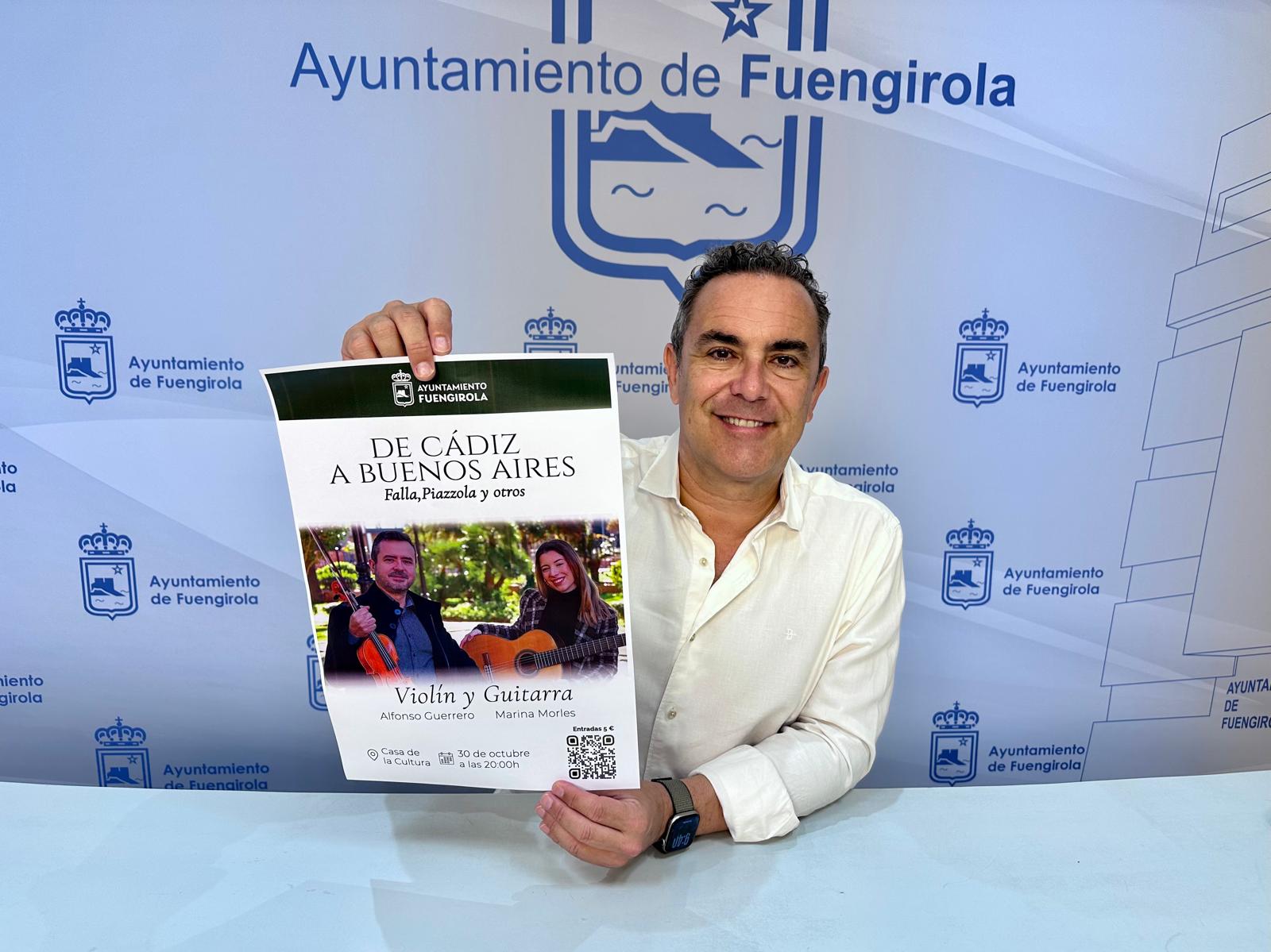 Un hombre sonriente sostiene un cartel de un recital de violín y guitarra ante un fondo con el escudo del Ayuntamiento de Fuengirola.
