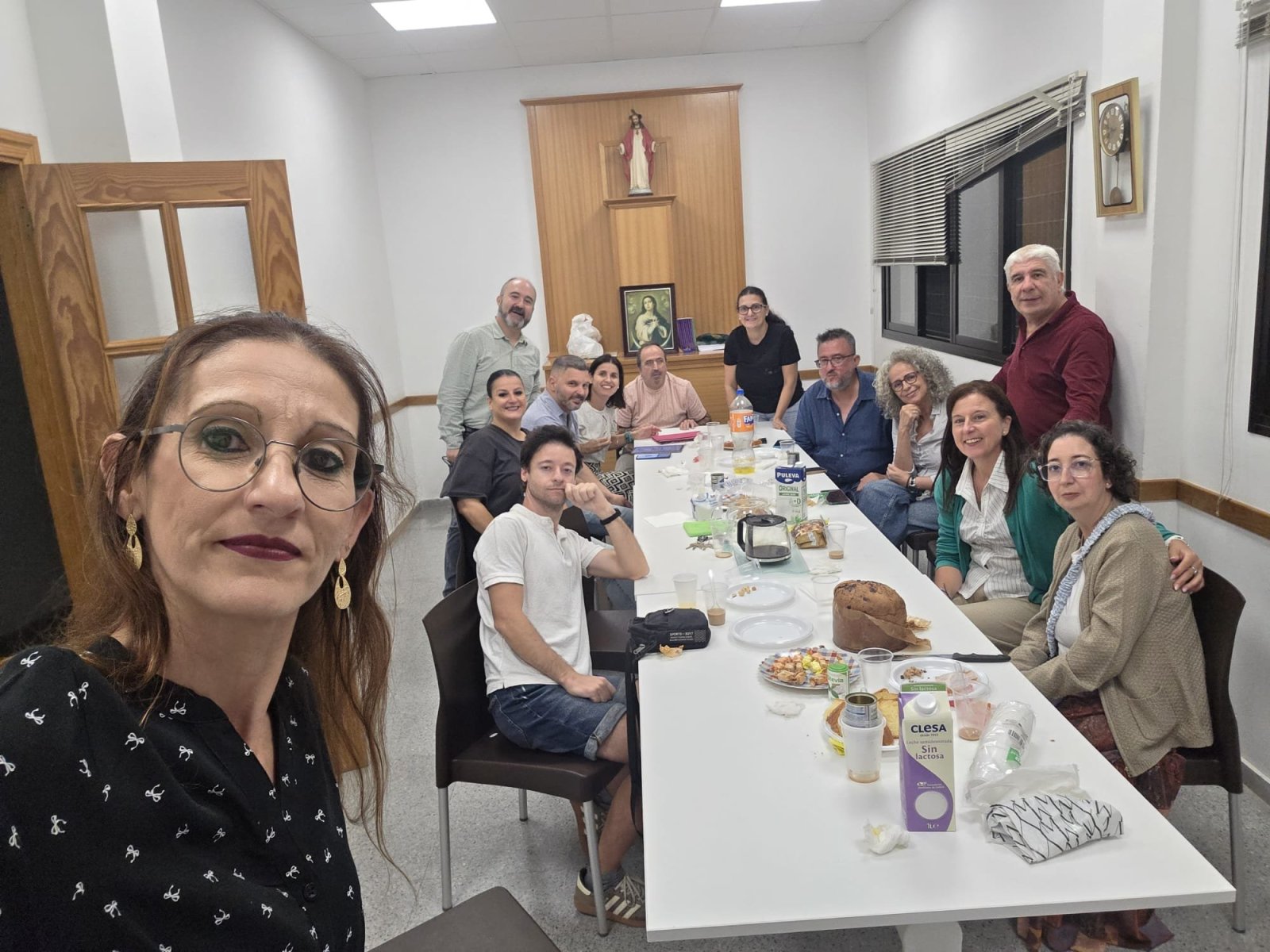 Una mujer con gafas se hace un selfi al frente de una sala con una mesa larga y un grupo de personas sentadas y de pie alrededor.