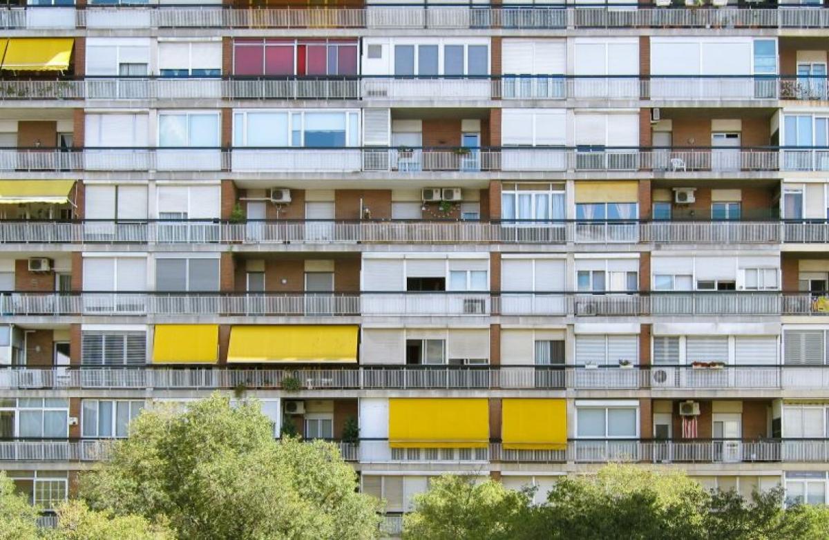 Fachada de un edificio alto de viviendas con balcones y toldos amarillos, parcialmente oculta por árboles en la parte inferior.