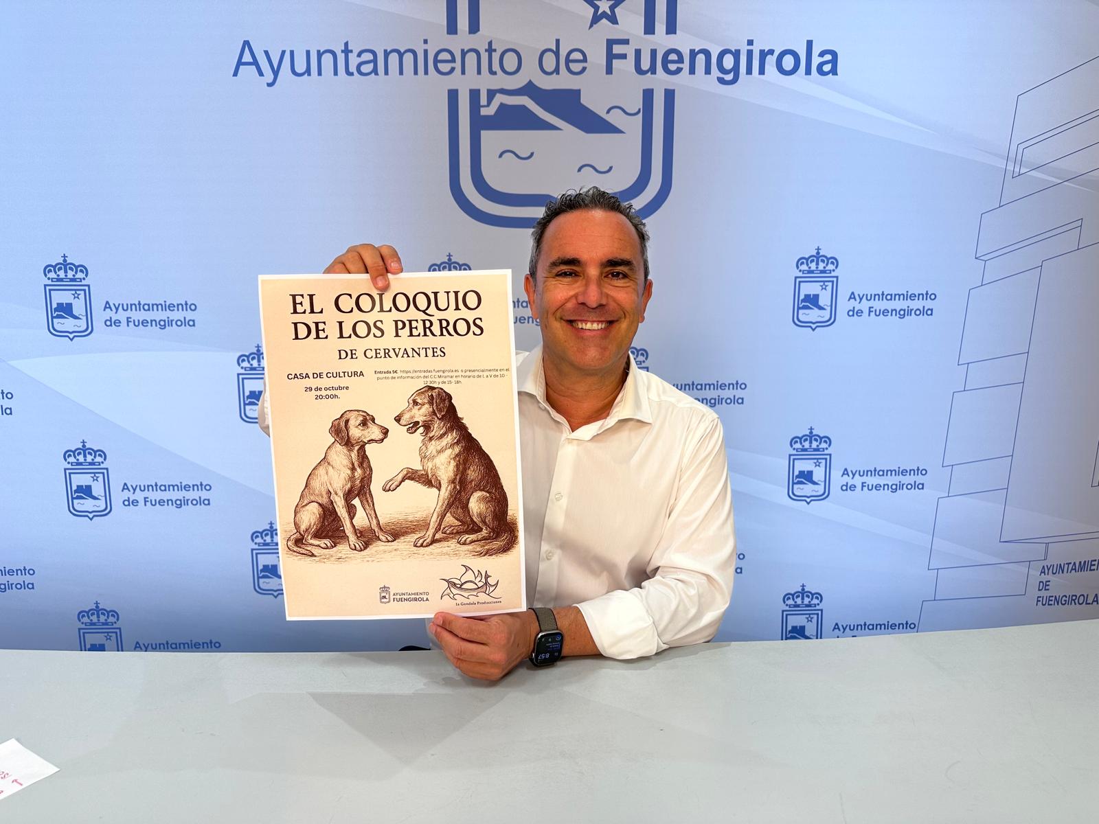 Un hombre sonriente sostiene un cartel de la obra El coloquio de los perros ante un fondo con el logotipo del Ayuntamiento de Fuengirola.