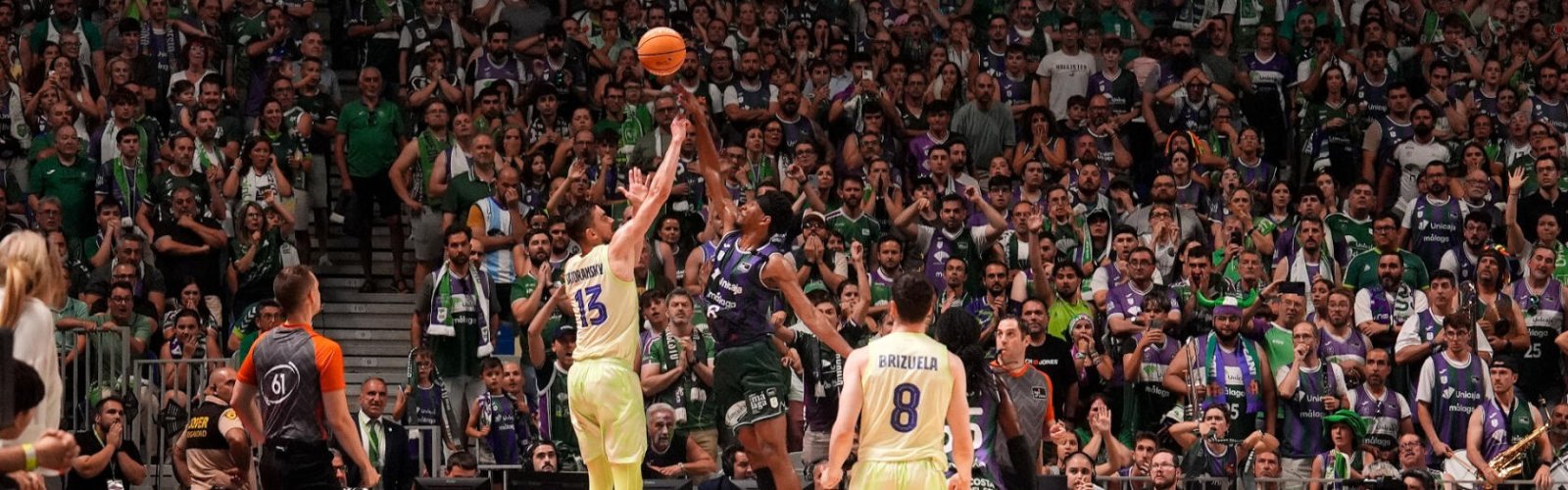Dos jugadores de baloncesto saltan junto al balón ante una grada llena de aficionados, con un árbitro y otros jugadores en la cancha.