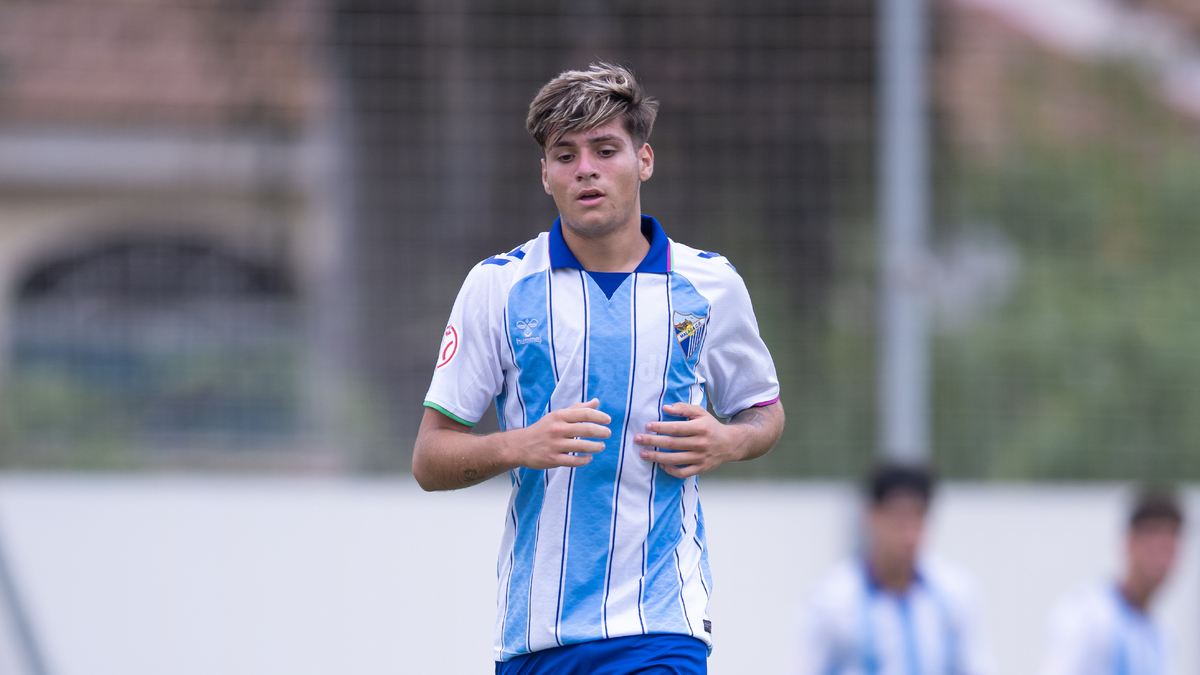 Un jugador de fútbol con camiseta de rayas azules y blancas camina sobre el césped, con otros jugadores desenfocados al fondo.