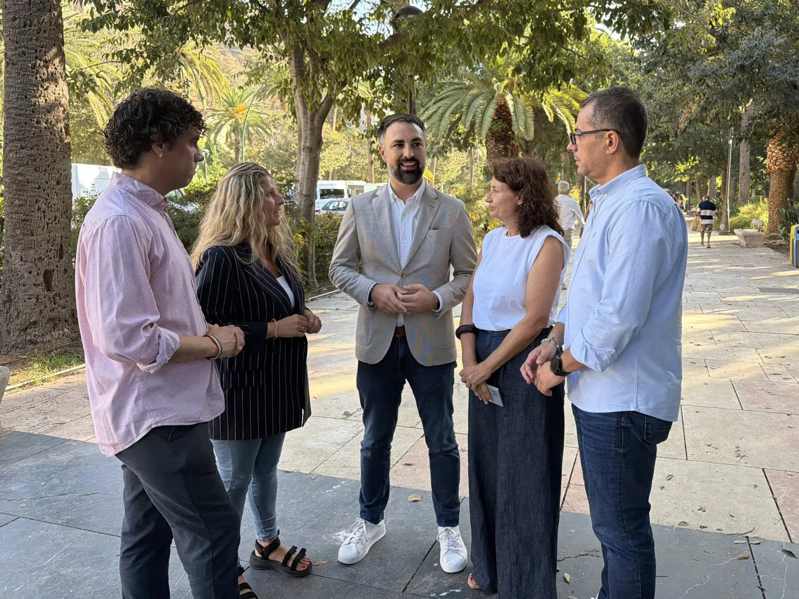 Cinco personas conversan de pie en un paseo pavimentado con árboles y palmeras al fondo.