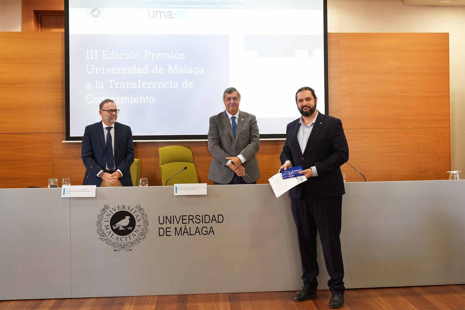 Tres hombres trajeados tras una mesa con el logotipo de la Universidad de Málaga, ante una pantalla que anuncia premios.