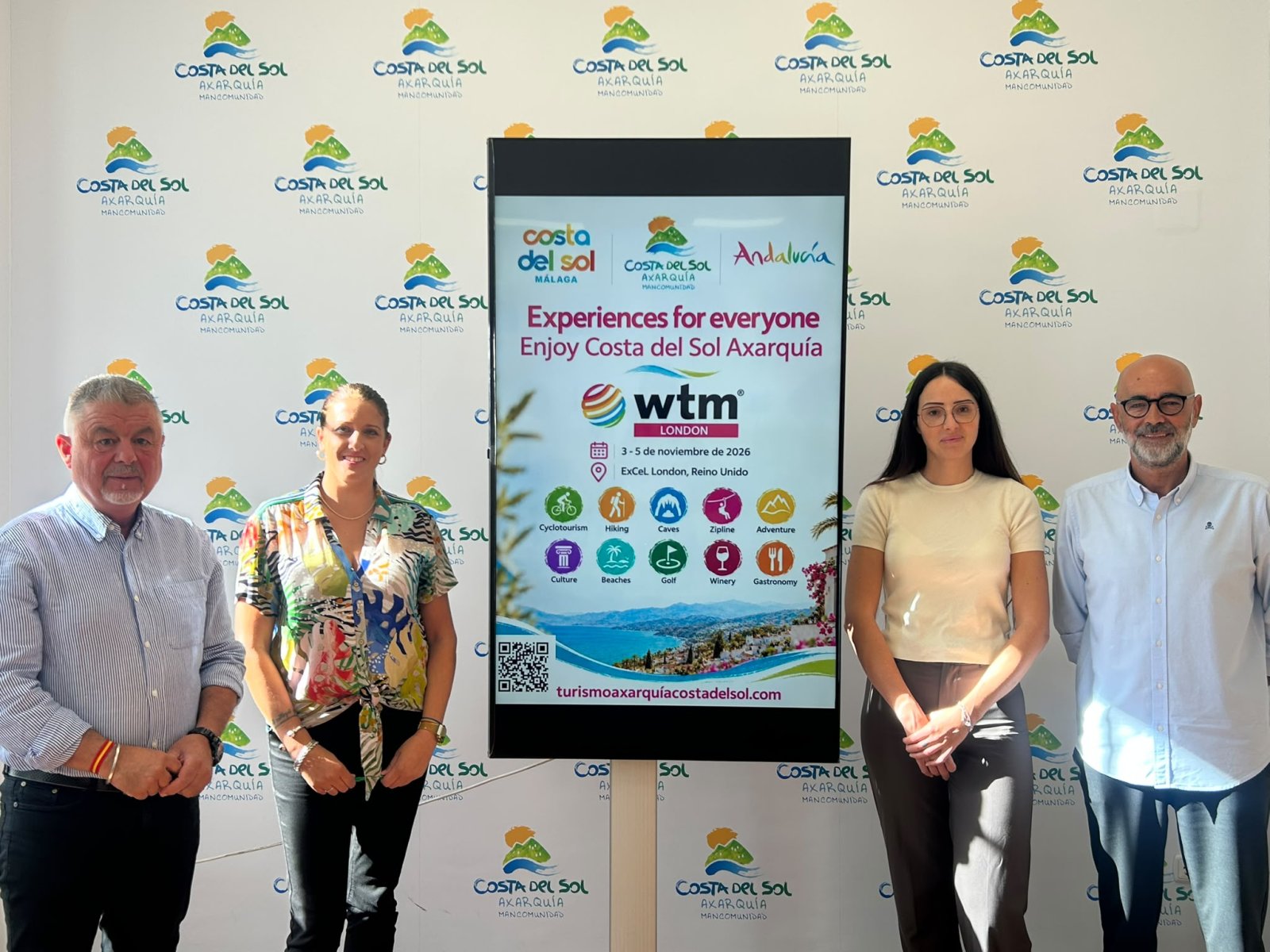 Cuatro personas de pie junto a una pantalla vertical con el cartel de la feria WTM de Londres, ante un fondo con logotipos.