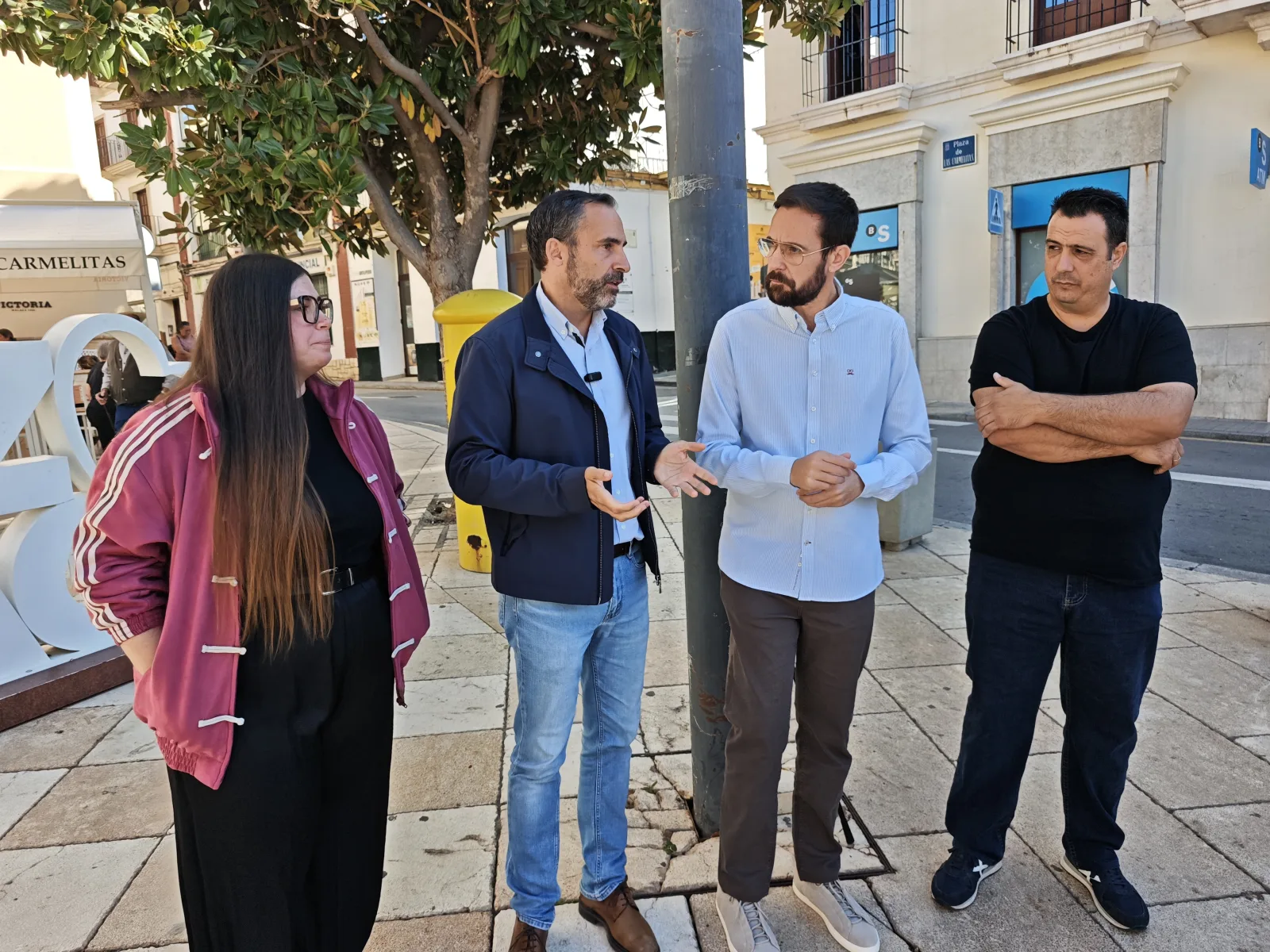 Cuatro personas de pie conversando en una acera junto a un poste, con edificios y un árbol al fondo.