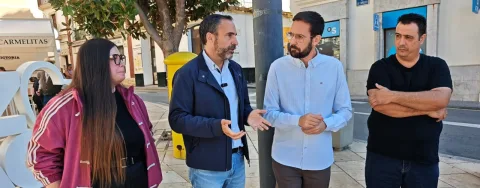 Cuatro personas conversan de pie en una acera junto a una farola y un buzón amarillo, con edificios y un toldo al fondo.