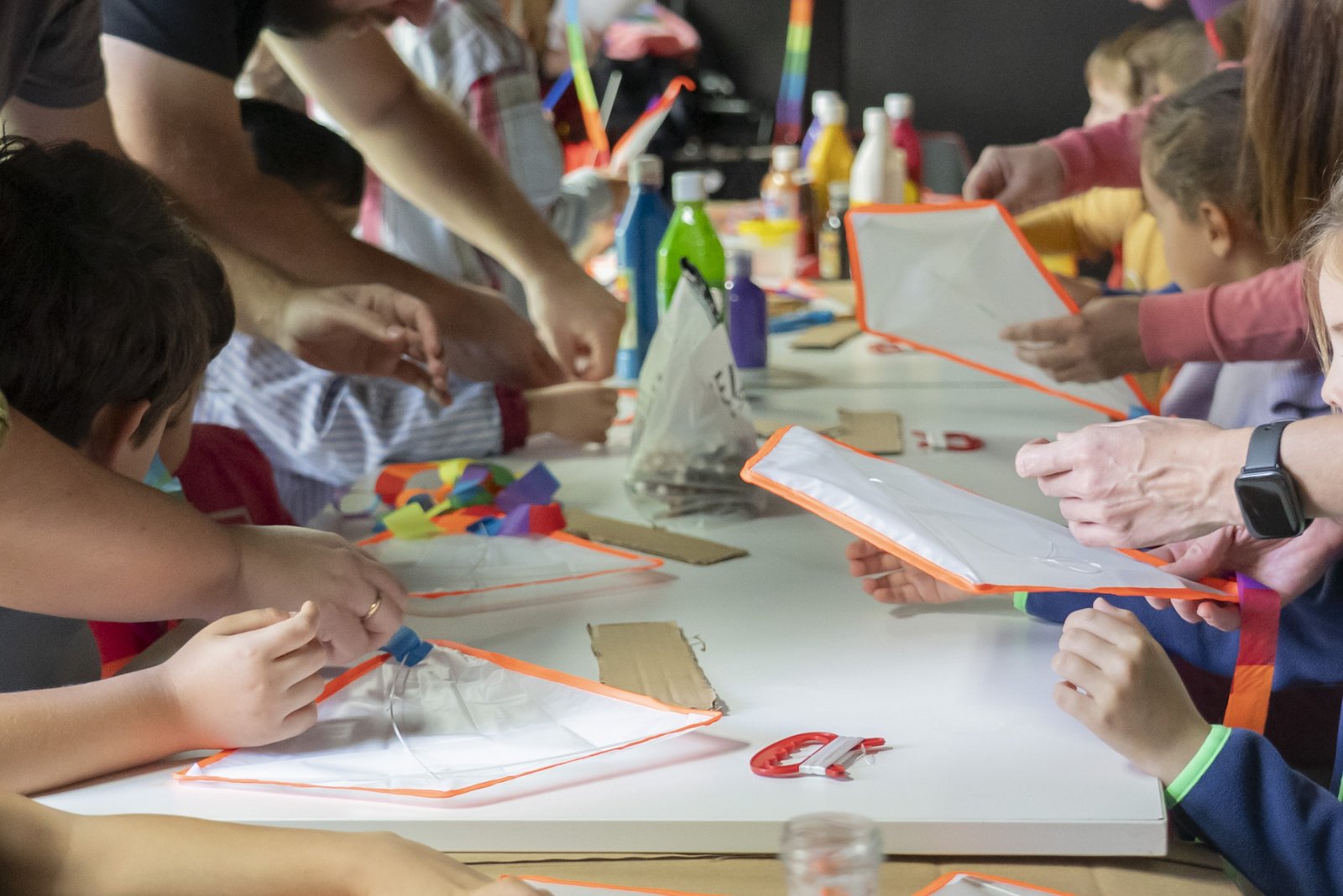 Niños y adultos con las manos en una mesa blanca manipulando fundas transparentes con borde naranja, con botes de pintura y papeles de colores al fondo.